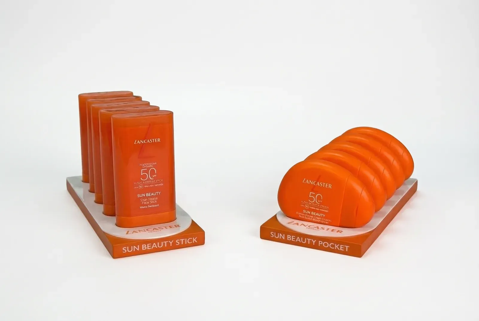 Posicionadores de sobremesa de Lancaster Sun Beauty: base de DM lacado naranja con el nombre de la variante serigrafiado e inserto de metacrilato blanco que aloja los sticks y las cremas de bolsillo