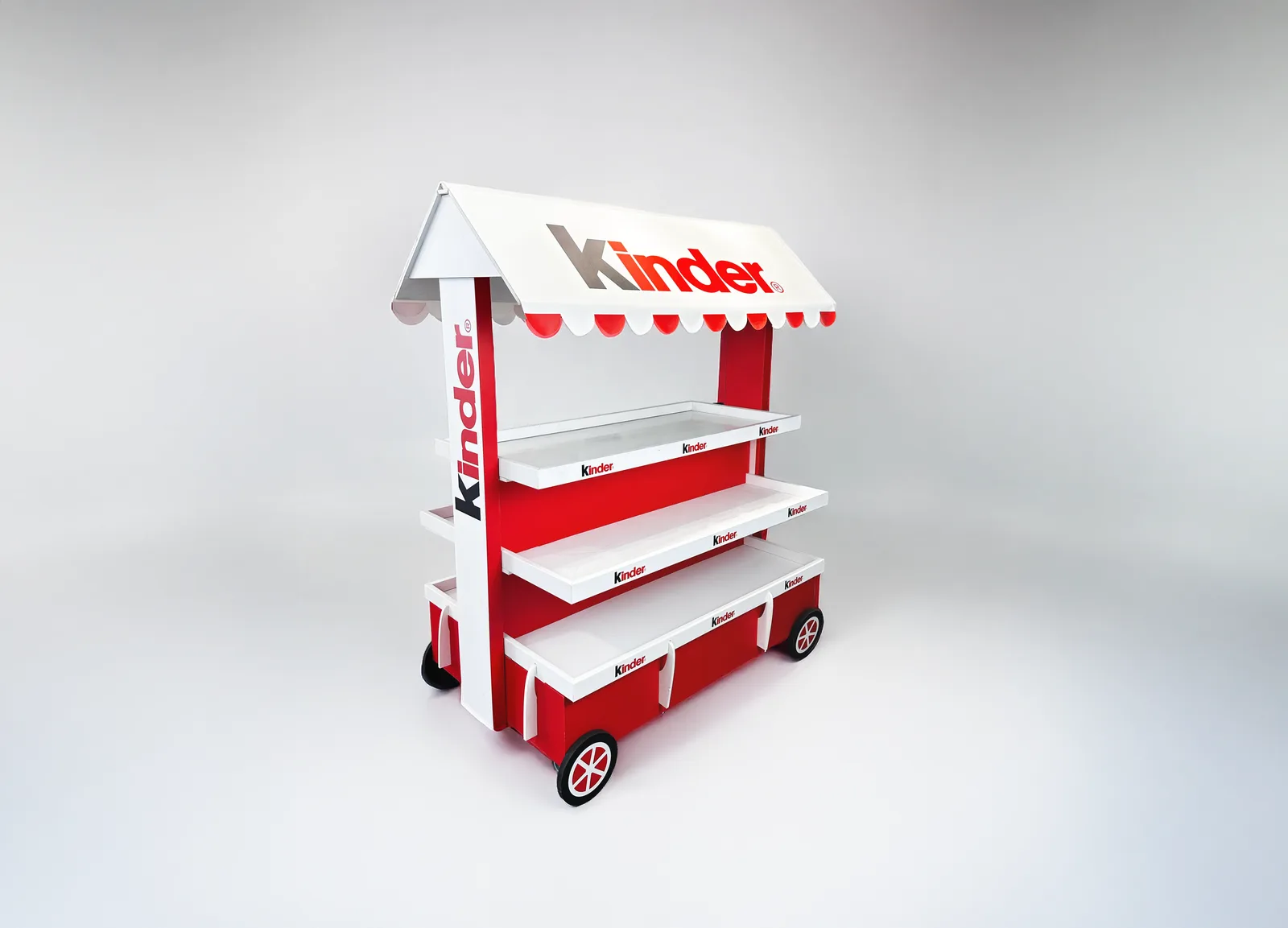 Expositor de suelo de Kinder con forma de carrito de mercado, toldo blanco a dos aguas con festón rojo, tres baldas blancas y ruedas de radios rojos, fotografiado vacío sobre fondo gris